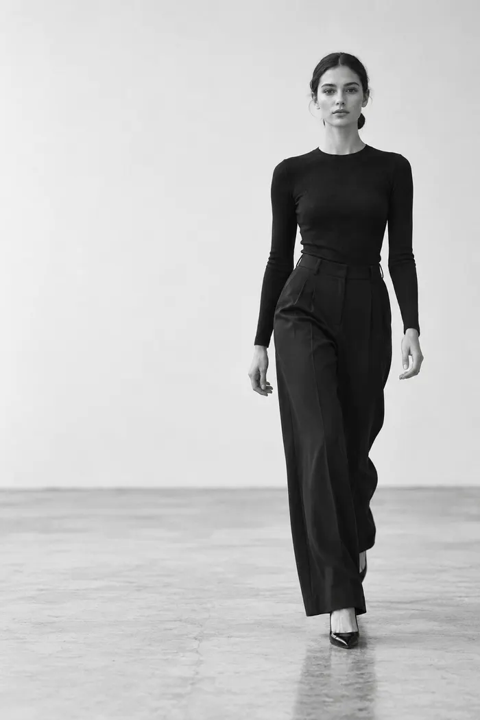 A model in a black top and wide trousers walking towards the camera in a white studio