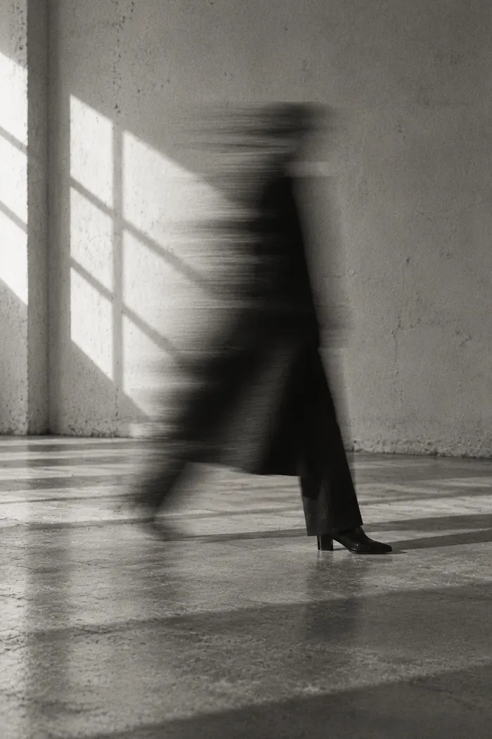 A motion-blurred figure in a long coat walking past a sunlit wall